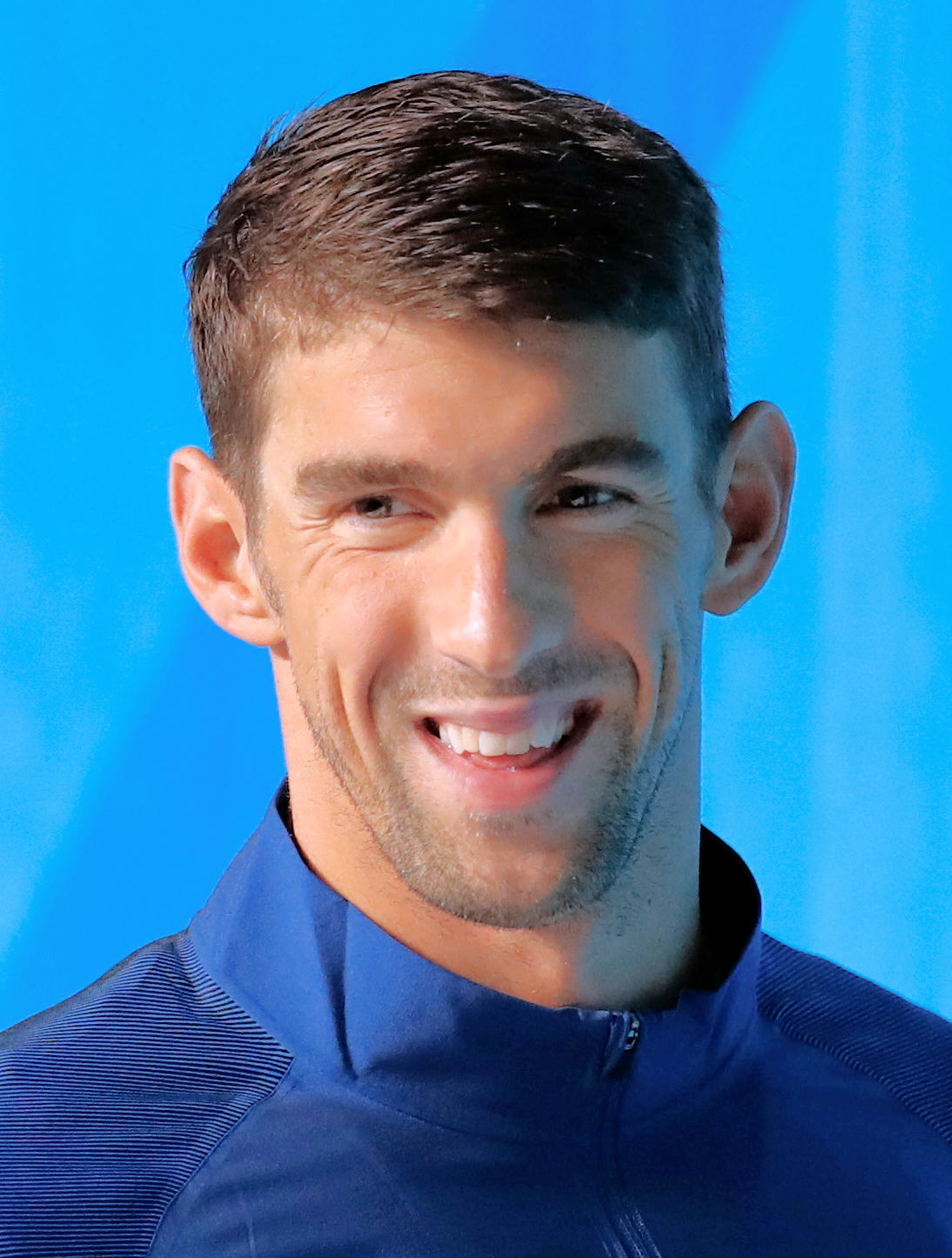 Close up picture of Olympic Swimmer Michael Phelps smiling while wearing a blue zip up hoodie infront of a light blue backdrop